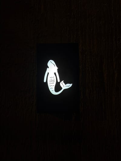 A vertically oriented photograph displays an illuminated white silhouette of a mermaid, centered within a black rectangular sign, mounted on a dark, textured wall in Ao Phai, Thailand. The mermaid figure faces right, featuring distinct hair, a human-like torso with arms, and a scaled fish tail adorned with wavy lines, terminating in a fluked end. The sign emits a bright white light, creating a strong contrast against its black frame and the surrounding wall. The wall exhibits a rough, uneven texture, characterized by vertical striations and variations in dark brown and black tones, consistent with stucco or a similar material. The environment is dimly lit, accentuating the glowing sign.