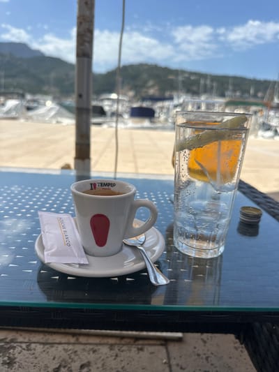 The image depicts a table setting in what appears to be El Port, Spain, based on the outdoor view and the text on the coffee cup. A glass of sparkling water with a lemon slice sits on a glass table, along with a small coffee cup with a spoon resting on the saucer. A packet of "Azúcar Blanco" (white sugar) is near the cup. The table is set outdoors with a view of a harbor with boats. The focus is on the drinks and coffee cup on the table. The image is taken at a slightly elevated angle.