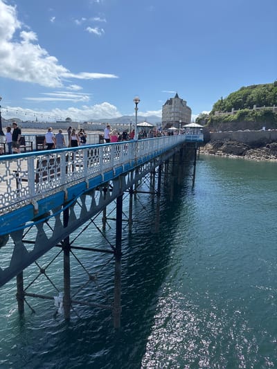 The image shows the Llandudno Pier, located in Llandudno, Wales, United Kingdom. It is a long pier extending out into the sea. People are walking along the pier, looking out at the water. The pier's structure is visible beneath the walkway, and there is a building at the end of the pier. The sea is calm and reflects the sunlight. The sky is blue with scattered clouds. A cliff face is visible on the left side of the image.