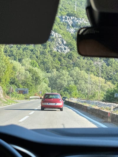 The image depicts a view from inside a vehicle traveling on a road, likely in or near Braćeni, Montenegro, given the road signs. A red hatchback car is in front, traveling in the same direction. The road has white lane markings and is flanked by a stone embankment on the right, with railway tracks and ballast. Beyond the embankment, a slope covered in dense green trees rises steeply, with rocky outcrops visible on the upper slopes. Overhead, power lines and poles are present. A signpost on the left side of the road indicates directions to "Skadarsko Jezero" and "Rijeka Crnojevića," confirming the location is in Montenegro, likely near Lake Skadar. The sun is shining, casting shadows on the road. The overall environment is mountainous and rural.