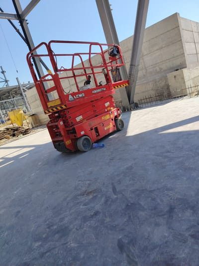 In Devnya, Bulgaria, a bright red aerial work platform (AWP), identified as a LGMG AS0607W, is positioned on a concrete surface under a clear blue sky. The AWP is angled, with its basket raised and a worker visible within it, appearing to be engaged in work. Structural steel beams, some bolted together, rise into the frame, indicating ongoing construction. A large concrete wall forms a backdrop, with rebar visible in the foreground, suggesting concrete foundation work. Other construction materials and debris, including a yellow plastic chute and wooden planks, are scattered in the background. The scene is illuminated by sunlight, casting shadows on the concrete ground. A blue plastic bottle lies on the ground near the AWP.