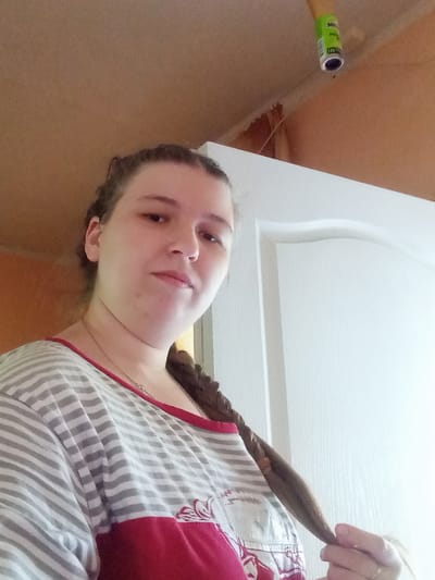 A medium shot captures a young woman indoors, facing forward and slightly to her left, with her right hand holding a strand of her braided hair. She wears a grey and white striped t-shirt with a red panel at the bottom featuring a white floral design. A thin necklace is visible around her neck. The background consists of a pale orange wall on the left, a white door on the right, and a beige ceiling above, where a bright green object, possibly a roll of tape or a small can, is affixed. The lighting is somewhat dim, with a strong light source to the right, creating a bright highlight on the door. The image is taken from a low angle, looking up towards the ceiling. This appears to be a residential interior, potentially in Kitskany, Moldova.