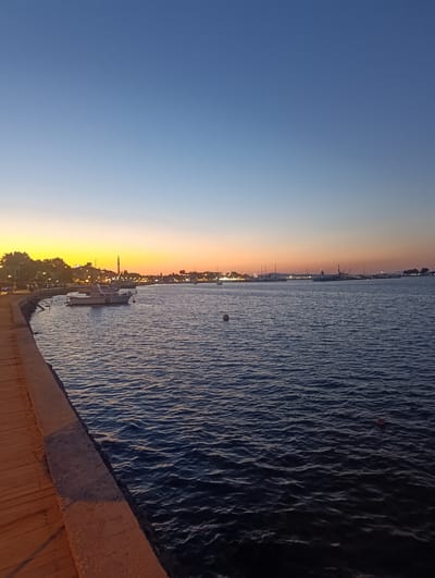 This is a landscape photograph taken at dusk, overlooking a harbor. The sky transitions from a deep blue at the top to a warm orange and yellow near the horizon, indicating sunset or sunrise. The water in the foreground is dark blue with gentle ripples, reflecting the colors of the sky. Along the left edge of the frame is a wooden walkway or pier with a stone edge.

In the middle ground, several boats are docked or anchored in the harbor. The closest boat is a white motor yacht. Further out, a line of buildings and structures is visible along the coastline, some with lights illuminated, suggesting a town or city. There are also masts of sailboats and possibly cranes, indicating a busy port.

The scene evokes a sense of calm and tranquility. The time of day is clearly evening or early morning. There are no people prominently visible, and the focus is on the natural beauty of the sky and water, combined with the man-made structures of the harbor. The location is identified as Urla, Turkey, which is consistent with coastal towns in the Aegean region. There is no visible text in the image.