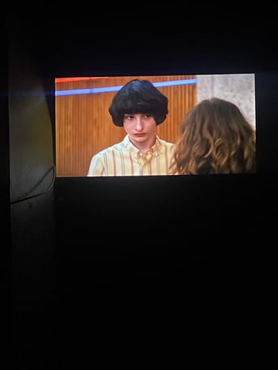 The image displays an indoor scene, set in Ksar-el-Kebir, Morocco, captured from a screen. A young male with dark, voluminous bowl-cut hair and a pale complexion occupies the left half of the frame, visible from the chest up. He is wearing a yellow and white vertically striped button-down shirt and looks forward with a neutral expression. Partially visible on the right is the back of another individual, distinguished by voluminous, wavy brown hair. The background consists of light brown wooden paneling, traversed by a horizontal red stripe positioned above a blue stripe. The surrounding areas of the photograph, particularly the lower half and left side, are dark, with a black cable partially visible along the far left edge.