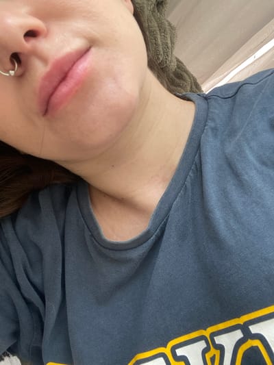 The image is a close-up, eye-level, medium shot of a person's face and upper chest, cropped from the nose down to the collarbone. The person has fair skin and is wearing a grey t-shirt with yellow and white lettering visible near the bottom of the frame. A silver septum ring is visible on the left side of the person's nose. Their lips are slightly parted and appear to be tinted pink. Behind the person's head, a textured, dark green towel or blanket is visible. The background is out of focus, showing a bright, white, vertical surface, possibly a wall or curtain. The lighting is soft and diffused. The image was taken indoors. The location of Romny, Ukraine, is not discernible from the visual content of the image.