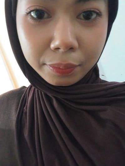 A close-up, head-and-shoulders shot of a young woman wearing a dark brown hijab. She is looking directly at the camera, with a neutral expression. Her skin is fair with a slightly dewy finish, and she is wearing subtle eye makeup and lip gloss. The background is out of focus, appearing to be a wall with a faint vertical line and a hint of yellow light source to the left. The lighting is soft, casting subtle shadows. The image was taken in Watampone, Indonesia.