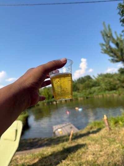 The image shows a close-up of a hand holding a clear plastic cup filled with a yellow, bubbly liquid, likely a beverage like beer or soda. The cup has ridged sides. The hand and cup are angled upwards against a bright blue sky with a few wispy clouds. In the background, out of focus, is a body of water, possibly a river or lake, lined with lush green trees. The sunlight suggests it is a sunny day, likely daytime. There are hints of a dock or wooden structure on the water, and some people are visible in the water as small, colorful figures, suggesting an outdoor recreational setting. The overall mood is relaxed and summery. The image was taken in Kamianka-Buzka, Ukraine.