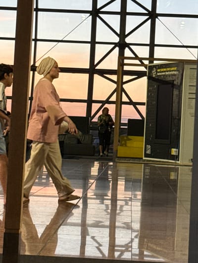 The image shows a hallway with a large glass ceiling with metal beams, through which the sunset is visible. A person in the foreground, wearing a light pink top and beige pants, walks from left to right. Their reflection is visible on the polished floor. To the right, a display panel with text and an image is mounted on a wooden frame. In the background, another person in a floral shirt and black shorts is standing near some yellow seating and a dark-colored sofa. This scene appears to be within Brussels Airport in Zaventem, Belgium, given the architectural style and the presence of airport-related signage.
