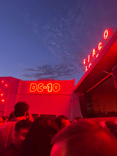 This is a medium shot of a crowd of people gathered outside DC-10, a nightclub in Sant Josep de sa Talaia, Spain, at dusk. The sky is a deep blue with some scattered clouds. The front of the building is illuminated by red neon lights, with a prominent "DC-10" sign in yellow neon. Above and to the right, a larger neon sign with yellow letters spelling "CIRCO" is visible. The crowd consists of multiple individuals, mostly their upper bodies and heads are visible, suggesting they are standing close together. The overall ambiance is that of a lively, outdoor event, likely a pre-party or waiting area before entering the venue. The red lighting creates a vibrant and energetic atmosphere.