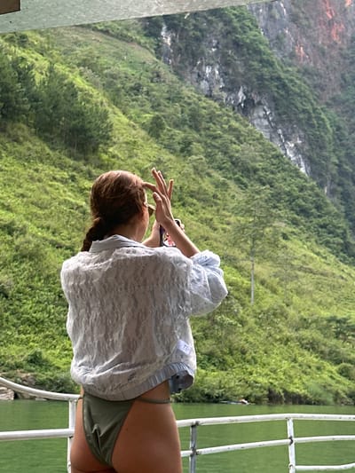 A woman with reddish-brown hair in a braid stands on a white metal structure in Đồng Văn Commune, Vietnam. She wears a light blue, lace-like top over a dark green bathing suit bottom. She appears to be using a smartphone to take a photo. The background is a steep, green, mountainous landscape with rocky sections.