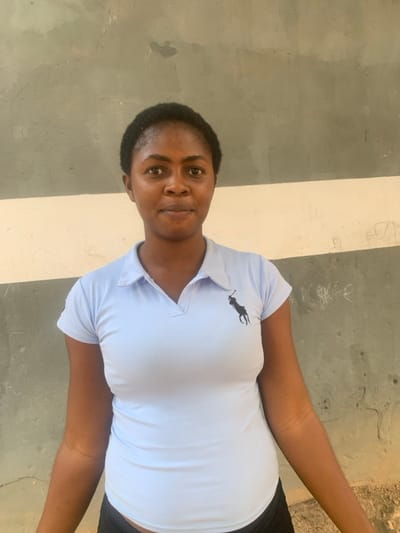 A woman stands near a striped wall, wearing a light blue polo shirt with a black logo. Her hair is short and dark. She looks directly at the camera with a neutral expression. Her dark pants are visible at her hip. Her arms are outstretched, and the surrounding environment appears to be an outdoor, possibly urban, setting.