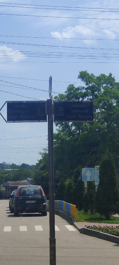 The image shows a street sign in the city of Kolomak, Ukraine, indicating directions to "ВУЛИЦЯ ГЕТЬМАНА ІВАНА МАЗЕПИ" (Hetman Ivan Mazepa Street). The sign is mounted on a pole with two black directional plates. The plate on the left points left, and the plate on the right points right. Both plates have the same text and a stylized symbol that appears to be a Cossack mace.
In the foreground, a black Chevrolet Aveo is parked, with its license plate visible as "AX 8852 OB". Behind the car, a colorful blue and yellow fence lines the side of the road. Further in the background, there are trees and buildings, including a sign with "РУСЛАНА" visible on a building. Traffic signs, including a directional arrow and a speed limit sign (30), are also present. The sky is blue with scattered clouds, and power lines are visible crossing the top of the frame. The scene appears to be on a bright, sunny day.