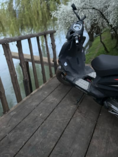 A dark-colored scooter is parked on a wooden bridge that appears to be over water. The bridge has gaps between the planks. On the left of the frame, there is vegetation and what appears to be a tree with white blossoms. The location is likely Brežice, Slovenia.
