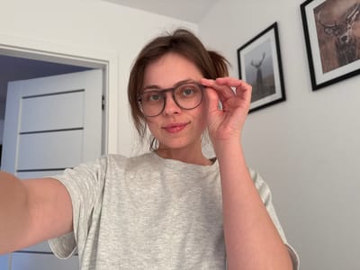 This is a portrait-oriented, slightly angled, selfie-style photograph of a young woman wearing glasses and a grey t-shirt. The image is upside down, so the woman's head is at the bottom of the frame and her feet (implied) would be at the top. She is looking directly at the camera with a neutral expression. Her hand is raised and she is touching the frame of her glasses with her index and thumb. Two framed pictures of deer hang on the wall behind her to the left. The room appears to be well-lit, suggesting it is daytime. The setting is likely a home or indoor space. There is no visible text in the image. The location context provided is Szklarska Poręba, Poland.