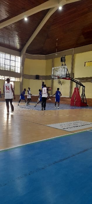 An indoor basketball court in Tudor ward, Kenya, is depicted during an active session involving approximately seven male individuals. The court floor features a wooden parquet surface with white and black markings, transitioning into a prominent blue section in the foreground, separated by a white line. On the right, a black basketball hoop support structure with red padding at its base holds a transparent rectangular backboard and a white net. A small oscillating fan is mounted on the support pole.
Multiple players are present. One player, wearing a white jersey with the number "7" visible on the back, stands centrally, holding an orange basketball near the hoop. Other players, dressed in either blue jerseys or white jerseys, are positioned around him, indicating active play. A player in a white jersey, numbered "14" on the back, is visible in the mid-ground on the left, walking away from the primary action.
The facility's walls are painted a light yellow, topped with a darker grey or brown horizontal trim line. Large rectangular windows with obscured glass panels are present on the left wall, providing natural light. The ceiling is high, vaulted, and constructed from dark wooden panels, supported by lighter-colored structural beams. Two illuminated spherical light fixtures are visible on the ceiling, contributing to the court's illumination.