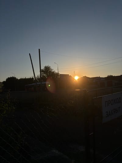 The image depicts a rural-urban transitional area in Новинки, Russia, at either sunrise or sunset, characterized by strong backlighting from a low sun. The bright, obscured solar disk is positioned above the horizon, slightly right of the frame's center, emanating significant lens flare with green, red, and orange concentric rings. The sky above is a clear, gradient blue, transitioning to warm yellow and orange hues near the horizon.
In the foreground, a dark, wire mesh fence (chain-link) runs diagonally from the lower-left to the center-right, casting deep shadows. To the far right, a solid dark metal gate post or barrier is visible, bearing a rectangular white sign with dark Cyrillic text. The visible portion of the text reads "ПРОХОД ЗАКРЫТ," which translates to "Passage Closed" or "Entry Closed." Dark, out-of-focus foliage occupies the extreme lower-left corner.
The midground features a road, mostly in shadow, traversed by a white and orange urban bus, partially silhouetted against the lighter sky. The number "15" is faintly visible on the bus's front, suggesting a route number. Two tall, slender, dark poles, possibly utility poles or temporary supports, lean slightly inward in the central-left area. Behind the road, a line of dark, dense trees creates a prominent silhouette against the horizon on the left.
On the right side of the midground, several low-rise buildings, likely residential houses or small commercial structures, are silhouetted, with some revealing peaked roofs. Overhead, power lines stretch horizontally across the frame. A faint "OZON" logo, indicative of an e-commerce platform, is discernible on a sign above one of the buildings on the mid-right. No individuals are explicitly visible in the scene.