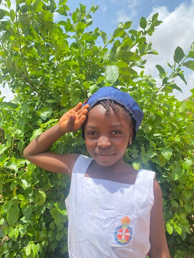 A young black girl wearing a blue hat and a white shirt with a "Girls Brigade" patch stands in front of a lush green bush on a partly sunny day. The location is likely Turu, Nigeria.