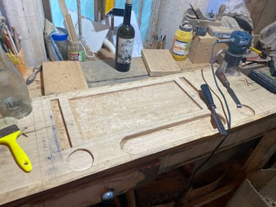In a workshop setting, likely in Negrești, Moldova, a wooden workbench is covered in sawdust and features various woodworking tools and materials. Prominently in the foreground is a large, unfinished wooden plank with carved indentations and a circular cutout. A yellow-handled brush and a bottle labeled "Mi Piace Strawberry Delight" are positioned to the left. To the right, a small electric router with its cord is visible, along with two chisels.
The background reveals a collection of items including a bottle of wine, a yellow bottle with the number "30" and text in Romanian, other tools, jars, and wooden blocks. The overall environment appears cluttered and actively used for woodworking. There are no people present in the image.