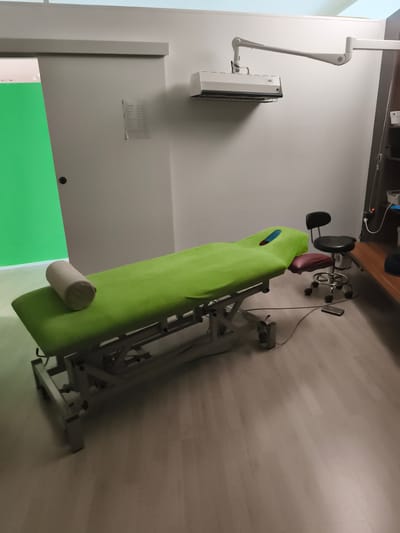 The image depicts a treatment room, likely a medical or therapeutic setting. A green-covered examination table with a beige bolster is centrally positioned. To the right of the table is a black adjustable stool with wheels and a maroon upholstered seat. In the background, a white wall is mounted with a light fixture on an articulated arm and an air conditioning unit. A tall, narrow green partition is visible on the left, suggesting a changing area or a backdrop. The room has light-colored wooden flooring. The overall impression is of a clean, functional space. There are no people present in the image. The provided context places this scene in Troisdorf, Germany.