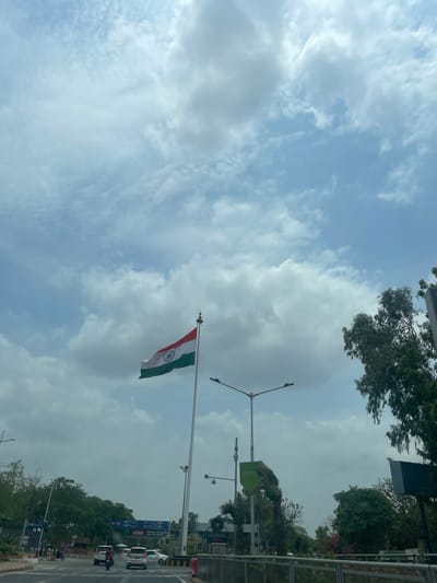 The image captures a view of the Indian flag flying high against a partly cloudy sky in Ahmedabad, India. The flag, with its distinct saffron, white, and green bands and the Ashoka Chakra in the center, is prominently displayed on a tall flagpole. Below the flag, a road with several vehicles, including cars and motorcycles, is visible. Streetlights and trees line the side of the road, suggesting an urban environment. Signage in the background indicates directions and possibly the name of a location. The overall scene depicts a bright, somewhat overcast day in a busy area of the city.