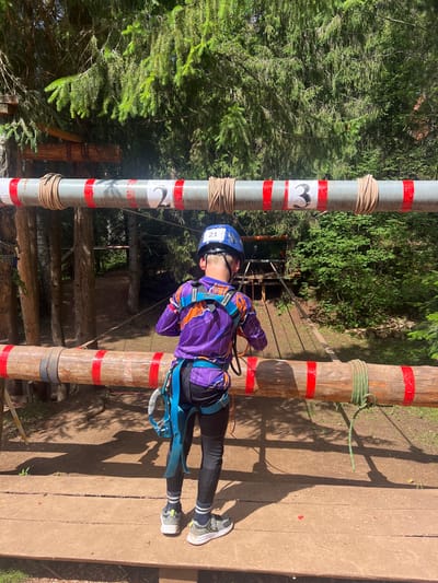 A young person, wearing a blue helmet and a purple and orange harness, is participating in an outdoor ropes course. They are clipped into a safety line and are positioned between two wooden poles that are part of the course. The poles are wrapped with rope and have red tape marking them at intervals. The young person is wearing dark leggings and athletic shoes. The background consists of lush green trees, indicating a forest setting. Sunlight is casting shadows on the wooden planks of the course, suggesting it is daytime. The scene appears to be in a recreational area designed for adventure activities. On one of the poles, there is a white label with the number "3" inside a black heart shape, and another label with the number "2". Another pole has the number "21" visible on a white sticker on the helmet. The overall impression is one of an active and adventurous outing.