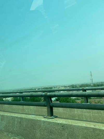 A roadside view features a concrete barrier with a metal railing. Beyond the railing, a hazy atmosphere obscures the distant landscape. A transmission tower can be seen among the buildings and trees. The sky is a light blue, blending with the haze, while a reflection is visible on the left side of the frame.
