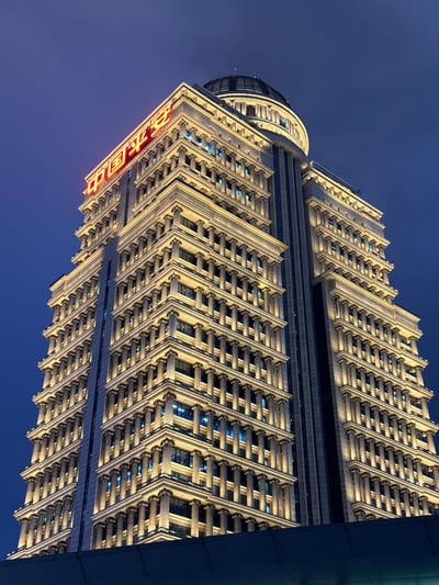 This media file showcases a striking, upward-angle view of a modern skyscraper at dusk in Pudong, China. The building's facade is illuminated by warm lights, highlighting its intricate architectural details. The design features repetitive, angled structures that create a sense of depth and complexity. The sky, a deep twilight blue, provides a dramatic backdrop.
On the side of the building, red neon Chinese characters are visible, adding a vibrant splash of color against the muted tones of the architecture and sky. The characters appear to spell out "中国银行," which translates to "Bank of China." The image captures the building from a low perspective, emphasizing its height and grand scale. The lighting suggests that it is either early morning or late evening, with the artificial lights of the city beginning to dominate the fading natural light. The overall mood is one of urban grandeur and modern architectural achievement.