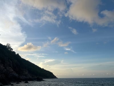 The image shows a coastal scene in Baan Chaloklum, Thailand. A steep, dark green, rocky hillside occupies the left side of the frame, with some trees at the top. The lower part of the hill transitions to a rocky beach with ocean water. The ocean, reflecting the sky, stretches horizontally across the bottom half of the image. The sky is predominantly blue, with scattered white clouds, some tinged with yellow. The horizon is visible in the distance. No people are visible.