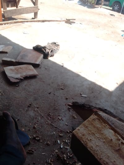 The image displays an outdoor, ground-level scene within the city of 0801712591, Nigeria. The immediate foreground features a dark-skinned human left foot, partially visible, wearing a dark-colored slipper or sandal. The ground consists of rough concrete or compacted dirt, heavily scattered with wood shavings, numerous light-colored peanut shells, and small debris. In the mid-ground, multiple pieces of cut wood are present: to the right, two rough-hewn timber planks with visible bark along one edge; to the left, several flat, irregular wooden offcuts, alongside a crumpled dark fabric item. A distinct diagonal shadow line separates a darker, shaded area from a brightly sunlit area of the ground. In the background, a wooden structure constructed from rough planks is partially visible on the left. On the far right, a portion of a green vehicle, possibly a bus or car, is discernible. The environment suggests an informal outdoor workspace or market setting under strong daylight.