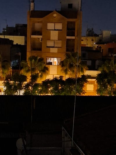 The image depicts a nighttime urban scene, seemingly in Xirivella, Spain. The prominent feature is a three-story, yellow-brown building with multiple rectangular windows, some illuminated from within. The building appears to have a gable-style roof with a circular detail. Palm trees are visible, partially obscuring the building. A dark, seemingly obscured area to the right of the image includes some structures and possibly a cable or wire spanning across. The sky is dark, indicating nighttime. Other buildings can be seen in the background.