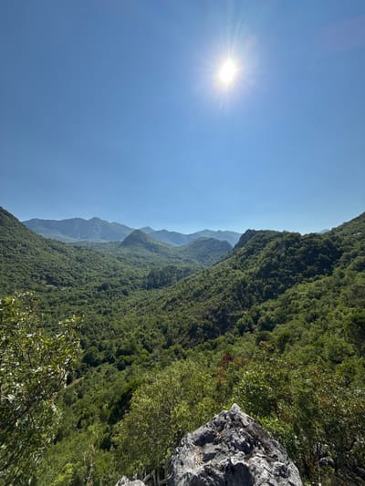 The image is a wide-angle, landscape photograph taken from a high vantage point, looking out over a lush, green valley filled with trees. The sky is a clear, vibrant blue with the sun shining brightly and prominently in the upper center of the frame, casting rays of light. In the foreground, a large, textured grey rock occupies the bottom right corner, partially obscuring some vegetation. The valley below is densely covered in a variety of green trees, with rolling hills and mountains stretching into the distance. The overall impression is one of a natural, scenic vista, likely in a mountainous or hilly region. The location is specified as Dupilo, Montenegro.