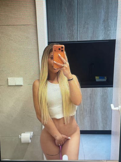 A blonde woman is taking a mirror selfie in a bathroom. She is wearing a white crop top and is holding her phone in her right hand, angled to capture her reflection. Her left hand is covering her lower body. The bathroom has light gray tiled walls, a light beige tiled wall with a light switch, and a dark gray cabinet. A roll of toilet paper is visible on a holder on the left wall. A black shelf with a yellow and blue object is mounted on the dark gray cabinet. A white door handle is visible on the right side of the image. The woman has blonde hair that reaches her shoulders.