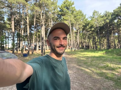 The image shows a man with a beard, wearing a green t-shirt and a tan baseball cap. He is facing the camera and appears to be taking a selfie. The background features a forest of tall, thin trees, possibly pine trees, with patches of grass and a light blue sky visible through the trees. The setting appears to be in a wooded area near Donji Štoj, Montenegro. In the background are vehicles and structures.