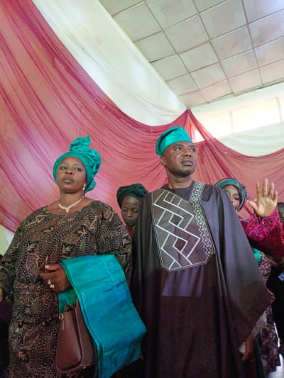 In Fagge D2, Nigeria, a medium shot captures a man and a woman standing indoors. The woman, wearing a brown patterned dress, a turquoise headwrap, and a diamond necklace, holds a turquoise shawl and a brown handbag. The man, beside her, wears a dark brown robe with geometric white embroidery on the chest and a matching turquoise hat. Behind them, draped maroon and white fabric serves as a backdrop. Other individuals are partially visible in the background. The setting suggests a special event or gathering.