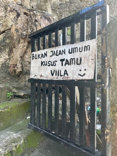 The image shows a black metal gate with a weathered wooden sign attached. The sign has black, handwritten text that reads "BUKAN JALAN UMUM KUSUS TAMU VIILA" and a smiley face. The gate is in front of a rocky embankment with some green vegetation and moss. To the left of the gate, there are moss-covered concrete steps leading up. The background appears to be an outdoor environment, possibly a hillside or a natural area, with some debris visible behind the gate. The location is identified as A R A, Indonesia.