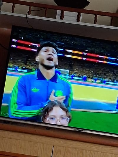 An indoor television screen displays a broadcast of a man in a blue and green athletic jacket with his hand on his chest, singing or speaking with his mouth open. Behind him, a crowd is visible in the background. In front of the television, a young person wearing glasses is looking up at the screen, their head partially obscuring the lower portion of the man on the screen. The television is placed on a wooden cabinet. There is a railing with wooden posts visible above the television. The lower portion of the image shows the wooden cabinet and a portion of the floor. The text "Z/RU" is visible on the lower left corner of the television screen.