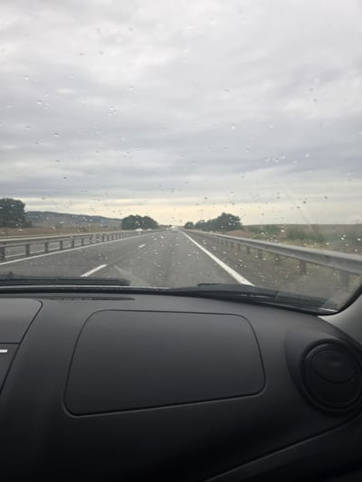 The image depicts a view from inside a vehicle, looking out through a rain-streaked windshield. The road ahead is visible, with a white line marking the lane. A metal guardrail runs along the side of the road, separating it from a grassy embankment and trees. The sky is overcast and grey, indicating a cloudy or rainy day. The time of day appears to be daytime, likely morning or afternoon, due to the ambient light. The interior of the vehicle is partially visible on the right side of the frame, showing a dark dashboard with a circular vent. The overall mood is somewhat somber, perhaps due to the weather. There is no visible text in the image. The location is Novyy Chirkey, Russia.