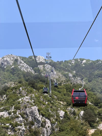 This media file shows a cable car system ascending a steep, rocky mountainside covered in dense green vegetation. The sky is a clear, bright blue, suggesting it is daytime and the weather is fair. The camera appears to be positioned inside one of the cable car cabins, looking out and down at the mountainous landscape and other cabins.

The primary objects are the cable car cabins, which are modern and appear to be made of metal and glass. One cabin, prominently in the foreground and slightly to the right, is red with black accents. It has a logo that reads "m-tel" in white text with a red stylized "m". Inside the cabin, faint text is visible on the windows. Other cabins, colored black with green accents and some with blue accents, are visible further along the cable line, ascending the mountain. These cabins are suspended from thick cables that stretch across the frame.

The scene is a rugged, natural environment. The mountains are characterized by large, light-colored rock formations interspersed with thick green trees and shrubs. The terrain is uneven and suggests a significant elevation change.

The activity taking place is the transportation of passengers via the cable car up the mountain. The perspective from within a cabin implies the viewer is a participant in this journey.

There is no explicit indication of people within the cabins, though their presence is implied by the function of the cable car. The overall mood is one of scenic travel and ascent, likely offering breathtaking views of the surrounding landscape. The clear sky and bright sunlight suggest a pleasant day for such an activity. The location is hinted to be Mirac, Montenegro, which is known for its mountainous terrain and scenic views.