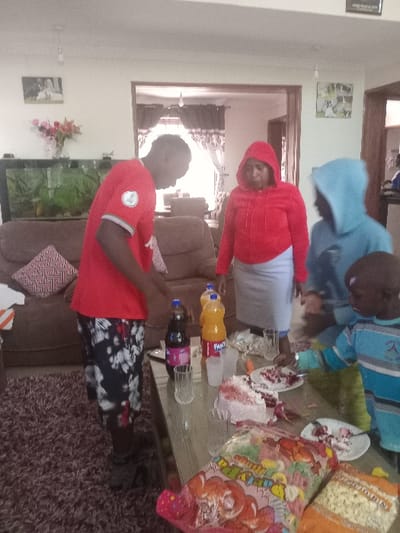 The image depicts an indoor scene within a residence located in Syokimau, Kenya, likely a living room or dining area. Four individuals are present around a glass-top table laden with food and beverages. The environment features light-colored walls, a dark brown shag carpet, and a large brown upholstered sofa in the background. A fish tank is visible behind the sofa, along with framed pictures and a vase containing flowers on the wall.

From left to right:
1.  A male individual, appearing to be a young adult, with dark skin, is positioned on the left side of the table. He is wearing a red short-sleeved t-shirt with a white emblem on the right sleeve, and black shorts with a white floral or leaf pattern. He is looking down towards the table.
2.  An adult female with dark skin is standing behind the table, centered in the frame. She is wearing a red hooded jacket and a light-colored skirt. Her gaze is directed towards the right side of the scene.
3.  A person, gender not definitively discernible, appearing to be an older child or young teenager with dark skin, is visible to the right of the adult female. This individual is wearing a light blue hooded sweatshirt and is positioned behind the table, interacting with the young child next to them.
4.  A young male child with dark skin is on the far right, reaching with his left hand towards a plate containing cake remnants on the table. He is wearing a long-sleeved shirt with horizontal blue and light blue stripes.

The glass-top table displays several items: a partially consumed pink or red cake with white frosting, multiple plates with cake remnants, two plastic bottles of soda (one dark, one orange Fanta), several clear glasses, and a white plastic cup. In the foreground on the table, a large clear plastic bag of colorful candies labeled "DELICIOUS" is visible, alongside a smaller orange bag, possibly containing popcorn. The overall scene suggests a casual gathering or celebration.