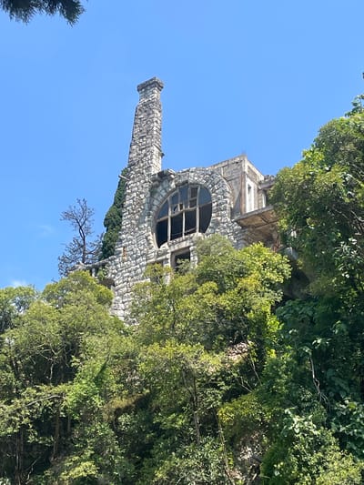 The image shows the abandoned and partially collapsed D.I. Gvazava Mansion, an architectural landmark located in Abaata, Abkhazia. The mansion is situated on a hillside amidst dense foliage. The structure is made of rough-hewn stone and features a prominent, tall chimney. A large, circular window with broken panes is visible on the facade. A balcony extends outwards, also appearing deteriorated. The surrounding environment is dominated by lush green trees and vegetation, with a clear blue sky overhead. There are no people visible in the image. The scene conveys a sense of decay and disuse.