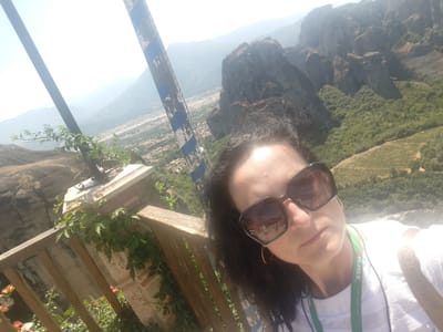 A woman is taking a selfie on a balcony overlooking a valley in Kalambaka, Greece. The woman has dark hair and is wearing large sunglasses and a white t-shirt. She is wearing a green lanyard around her neck. In the background, there are dramatic rock formations with trees and buildings at their base. There is a wooden railing in the foreground. A striped pole is visible on the left side of the image. The sky is bright and hazy.