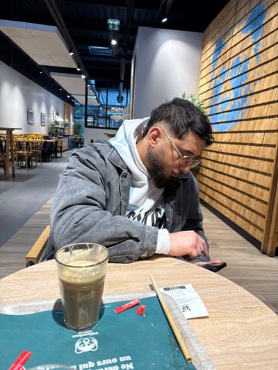 The image depicts an indoor scene within a cafe, identified as being in Reichstett, France. A male subject, appearing to be in his twenties or early thirties, is seated at a light-colored wooden table. He has short dark hair with faded sides, a beard, and is wearing round, light-colored framed glasses. His attire consists of a white hooded sweatshirt with a graphic design and a dark grey, stone-washed denim jacket. His head is tilted downwards as he actively engages with a black smartphone, holding it in his right hand.

On the table in the foreground, directly in front of the subject, rests a clear glass containing a dark, opaque liquid with a lighter foam layer on top, consistent with a coffee or smoothie drink. The glass sits on a teal-colored placemat or tray liner featuring a white logo of a bear-like figure and French text. Beside the glass, a broken red "Canderel" sweetener packet and another intact red packet are visible. To the right of the placemat, a light-colored paper straw lies parallel to a crumpled white receipt.

The background reveals a modern cafe interior. To the subject's right, a wall is adorned with horizontal wooden slats, featuring a large, stylized blue map design. To the subject's left and further back, additional cafe seating consisting of wooden tables and dark chairs with yellow accents is visible. The ceiling is dark with exposed utility elements and track lighting fixtures. Large windows or glass partitions are discernible further in the background, suggesting natural light sources or views to an exterior area.