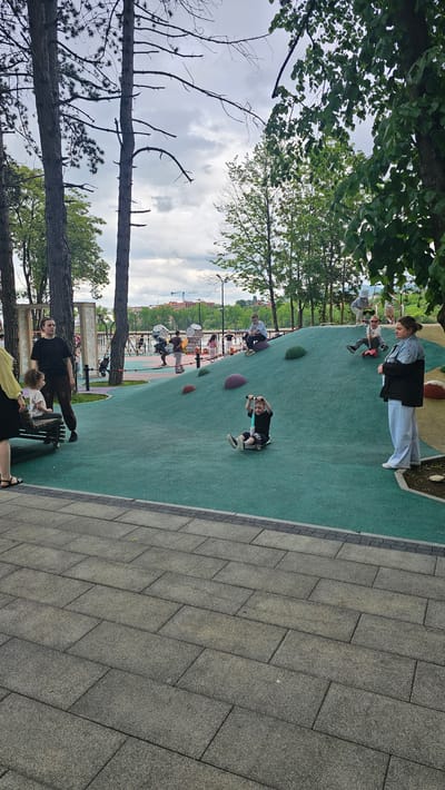 A group of people are at a park in Alikonovka, Russia. The park features a large, green, undulating play structure with several children playing on it. One child is riding a scooter down the hill, while others are sitting or playing on the structure. Adults are standing around watching the children. The park is surrounded by trees and a cloudy sky. In the background, there is a body of water and buildings.