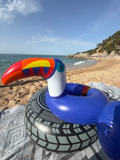 The image depicts a beach scene in Lloret de Mar, Spain, with a bright blue inflatable toucan pool float and a black inflatable tire tube resting on a grey and white striped beach towel spread on the sand. The toucan float, with its vibrant red, orange, yellow, and blue beak, is positioned in the foreground, its blue body forming a life preserver. Behind the floats, the sandy beach extends towards the ocean, where gentle waves are breaking. In the background, rocky outcrops covered in green vegetation line the coast, and a section of the beach is visible with several sunbathers and umbrellas. The sky is a clear, pale blue with a few wispy white clouds.