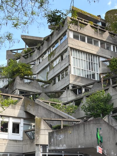 A photograph, rotated approximately 90 degrees clockwise, captures a striking brutalist-style concrete building in Ivry-sur-Seine, France, against a clear blue sky. The multi-story structure is defined by its complex, angular geometry, featuring numerous interconnected concrete beams, triangular sections, and recessed balconies. Lush green vegetation, including ivy and various bushes and small trees, extensively covers parts of the concrete facade and grows from planters within the building's design, creating a vibrant contrast with the raw grey material.

On the far left, a cylindrical, ivy-covered tower-like structure rises. The building is interspersed with numerous windows of varying sizes, predominantly featuring white frames, some of which are large and multi-paned. Tree branches with green foliage partially frame the scene from the lower left and upper right foreground.

Notable details include a green illuminated sign, possibly an arrow or directional symbol, attached to a concrete beam, alongside a red sign displaying "24H" vertically in white text. Faint graffiti markings are visible on the concrete in several areas. The bright daylight and clear sky suggest the photograph was taken during the day under fair weather conditions. The scene is static, with no people or discernible activity present.