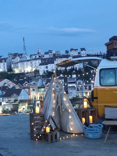 The image shows a nighttime scene in what appears to be Lang Biang - Đà Lạt, Vietnam. A teepee tent decorated with string lights and a dream catcher is on a rooftop, with wooden crates holding candles nearby. A yellow and white van is partially visible to the right, its open top adding to the ambiance. In the background, a hillside is covered with closely built houses and buildings, also illuminated. The sky is a dusky blue. There are no people visible in the immediate view.