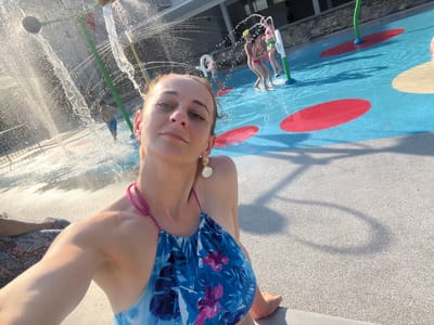 A woman is taking a selfie at a splash pad. She is wearing a blue and pink floral swimsuit and large, white, shell-shaped earrings. Behind her, children play in the water. One child in a pink swimsuit is being held by another child in a green swimsuit. Another child in blue shorts is running through a water spray. The splash pad has blue surfaces with red and cream-colored circular markings on the ground. Water jets are spraying from green poles and overhead structures. The sun is shining brightly, casting shadows on the ground. The environment appears to be an outdoor recreational area, possibly at a public park or resort.
