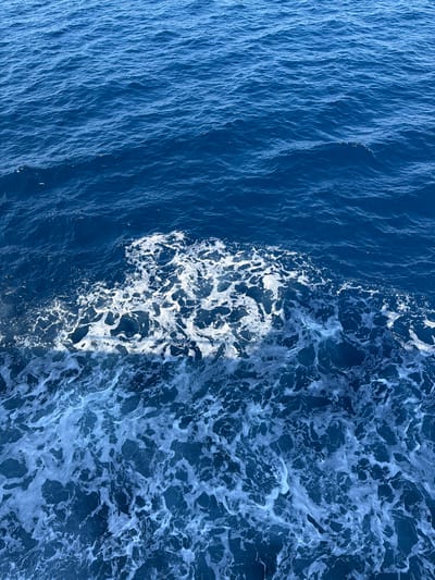 The image depicts the open sea with gentle waves and white foam. The water is a deep blue, and the sunlight reflects off the surface, creating a shimmering effect. The foam appears to be a wake, suggesting that a boat or ship has recently passed through the area. The horizon is not visible in the frame. The scene is likely a location in the sea off the coast of Qala, Malta.