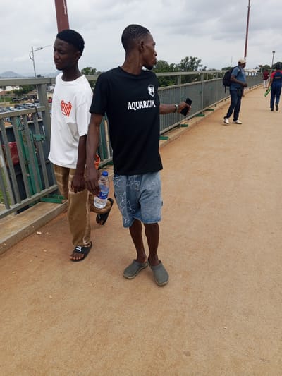 The image depicts two young Black men walking on a pedestrian bridge in Sauka, Nigeria. The man on the left wears a white t-shirt with red text and tan pants. He is wearing black sandals with a white "3" logo. The man on the right wears a black t-shirt with "AQUARIUM" printed in white text and denim shorts with white star patterns. He is wearing grey slip-on shoes. The man on the right is holding a water bottle in his left hand and a smartphone in his right hand. The bridge has a light brown paved surface and is bordered by metal railings. In the background, there are trees and some buildings, suggesting an urban or semi-urban environment. Further in the distance, a mountain is visible under a cloudy sky. The weather appears overcast, indicating a possibility of rain or a generally dull day. The time of day is not explicitly clear but the lighting suggests it is daytime. The overall mood is casual, with the two individuals seemingly engaged in conversation or simply walking together. There are other people visible on the bridge in the background, one of whom is wearing a uniform with the text "NSCDC" on it. The scene is a public space, likely a bridge over a road or a river, enabling pedestrian movement.