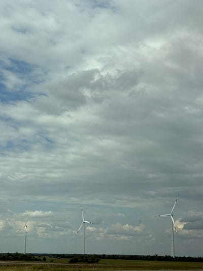 The image depicts a wide shot of a cloudy sky over a flat landscape with three wind turbines. The wind turbines are white, tall structures with three blades each, positioned at intervals across the horizon. The foreground shows a grassy field with a body of water or wet land. In the background, a line of trees and vegetation separates the field from the turbines. The sky is filled with dramatic grey and white clouds, with patches of blue sky visible. The scene appears to be a rural or semi-rural setting, likely a wind farm. The location is specified as Mayak, Russia.